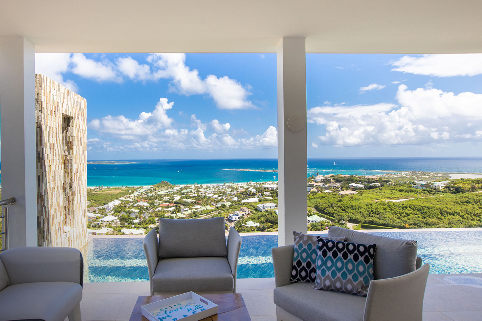 Elegant patio lounge with modern armchairs and infinity pool, showcasing a panoramic view of the vibrant blue coastline and scattered sailboats below.