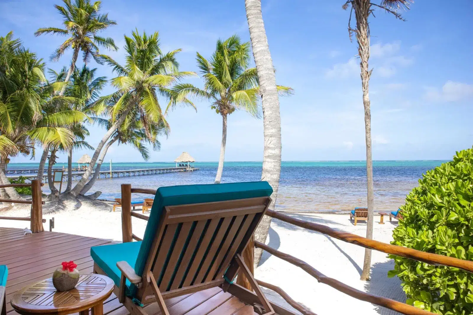 Oceanfront deck with wooden lounge chairs under swaying palm trees overlooking the Caribbean Sea in Belize, with a dock extending into the turquoise water.