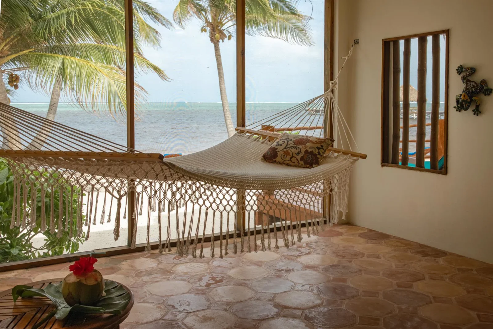 Indoor hammock facing large windows with a beachfront view in Belize, surrounded by palm trees and white sand, with a fresh coconut drink on a nearby table.