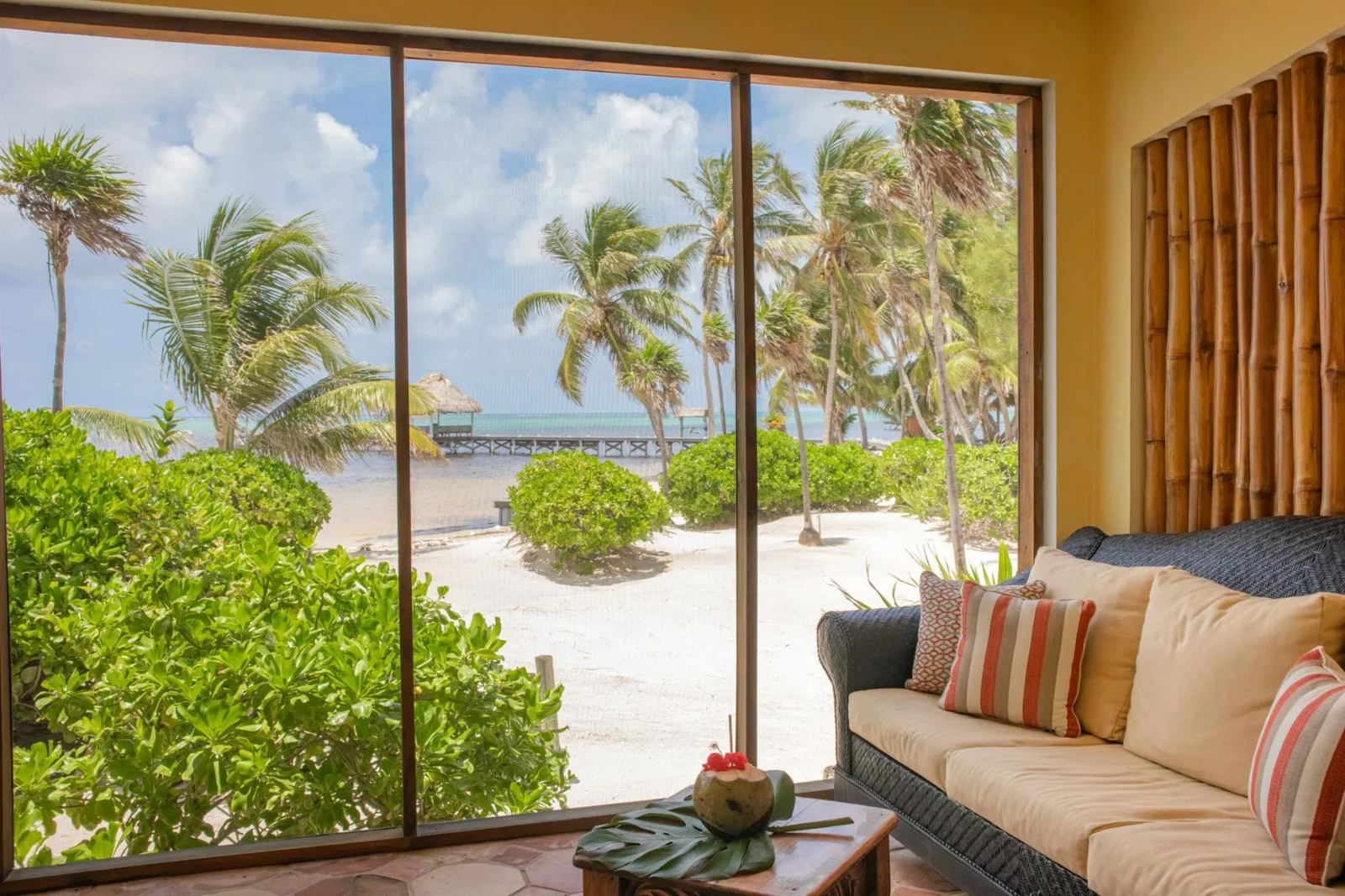 Scenic view through a villa window in Belize showing lush tropical greenery, white sand, and a private dock over the Caribbean Sea, with a cozy couch in the foreground.