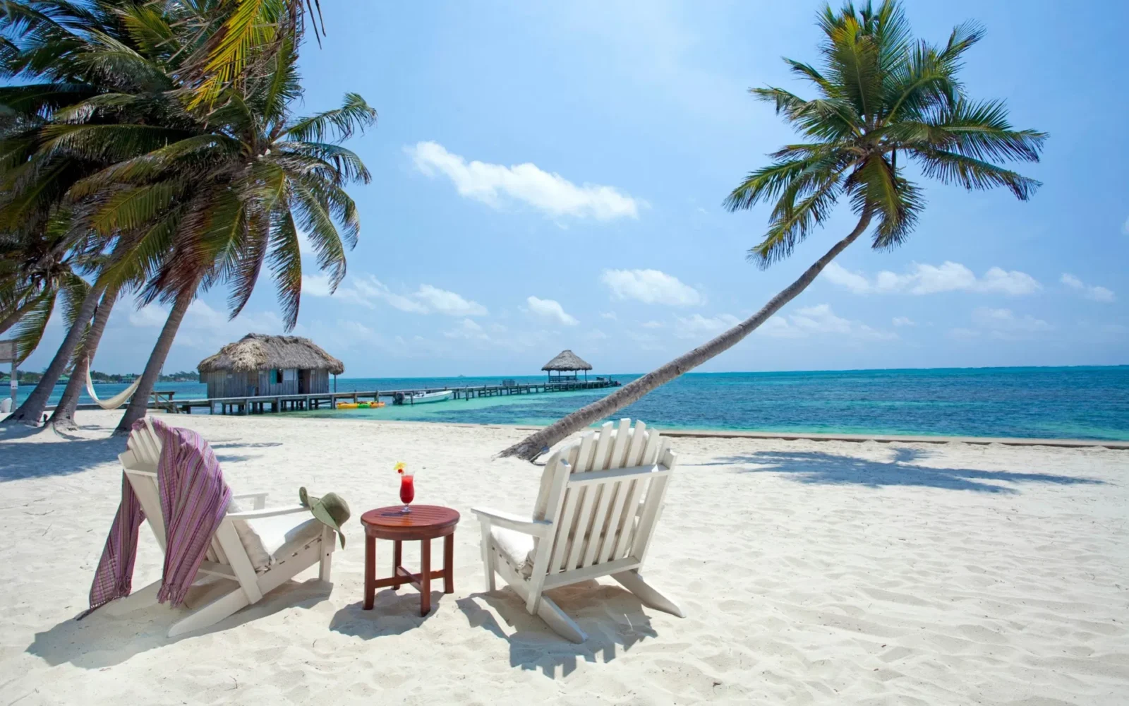 Two Adirondack chairs under palm trees on a white sandy beach in Belize, facing the ocean with a tropical drink on a table and a private dock in the distance.